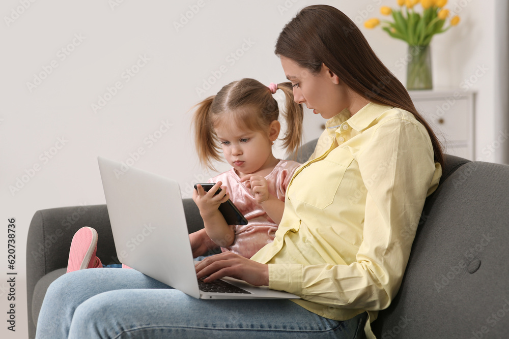 Mother working remotely on laptop while her daughter playing with smartphone at home Mother working remotely on laptop while her daughter playing with smartphone at home