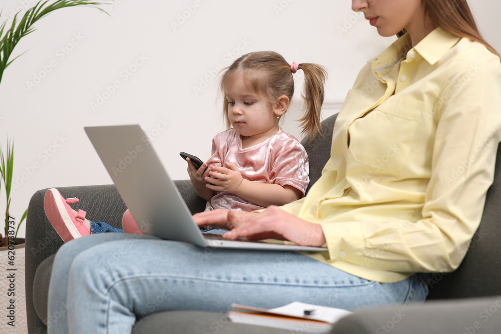 Mother working remotely on laptop while her daughter playing with smartphone at home, closeup