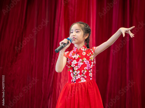 The Asian kid girl sing a song on stage at her school activity day, dress in Qipao style, red curtain background