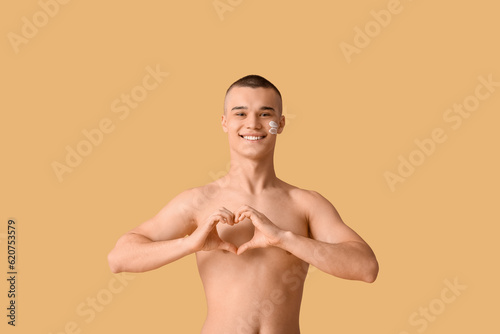 Happy young man with sunscreen cream on his face making heart gesture against orange background