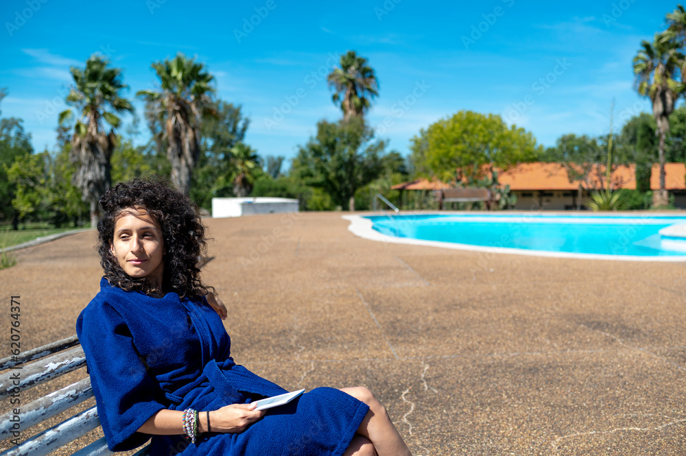 Woman relaxing sitting on a bench outdoors near swimming pool.