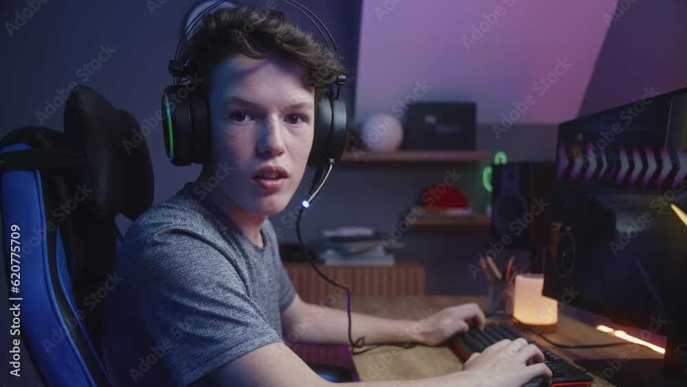 Young gamer in headphones plays on computer at home. Boy turns, looks and talks into the camera. Car racing video game displayed on PC monitor. Schoolboy spending leisure time. Dolly shot. Side view. Young gamer in headphones plays on computer at home. Boy turns, looks and talks into the camera. Car racing video game displayed on PC monitor. Schoolboy spending leisure time. Dolly shot. Side view.