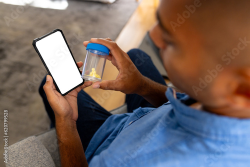 Biracial man holding medication making video call at home using smartphone with copy space on screen
