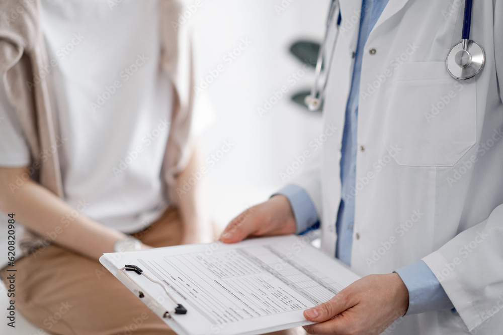 Doctor and patient discussing perfect health exam results. Friendly physician keeping with hands clipboard with medical papers near young woman. Medicine concept Doctor and patient discussing perfect health exam results. Friendly physician keeping with hands clipboard with medical papers near young woman. Medicine concept