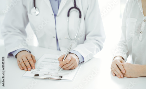 Doctor and patient sitting near of each other at the desk in clinic. The focus is on female physician's hands filling up the medication history record form or checklist, close up. Medicine concept