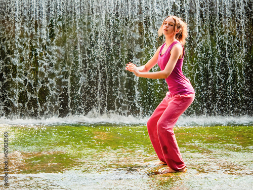 Young teenager girl doing body exercises in a small river, water fall out of focus in the background. Workout in outdoor area. Fresh energy concept. Healthy lifestyle. Stretches routine. Yoga style