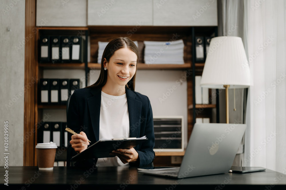 Confident business expert attractive smiling young woman typing laptop ang holding digital tablet on desk in office.. Confident business expert attractive smiling young woman typing laptop ang holding digital tablet on desk in office..