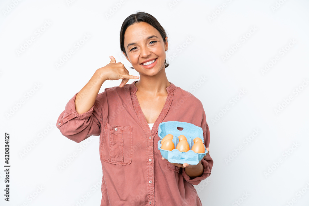 Young hispanic woman holding eggs isolated on white background making phone gesture. Call me back sign Young hispanic woman holding eggs isolated on white background making phone gesture. Call me back sign