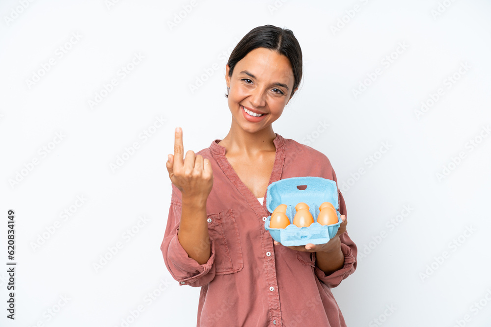 Young hispanic woman holding eggs isolated on white background doing coming gesture Young hispanic woman holding eggs isolated on white background doing coming gesture