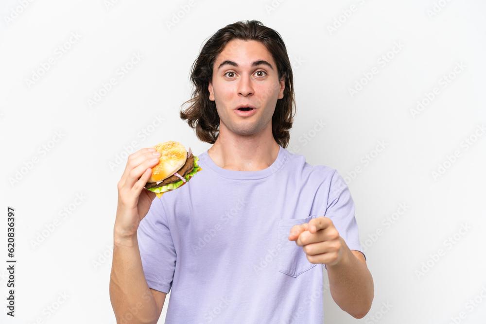 Young handsome man holding a burger isolated on white background surprised and pointing front Young handsome man holding a burger isolated on white background surprised and pointing front