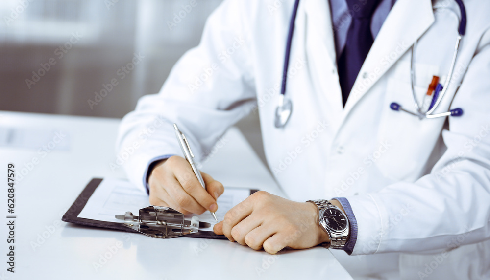 Unknown male doctor sitting and working with clipboard of medication history record in clinic at his working place, close-up. Young physician at work. Perfect medical service, medicine concept Unknown male doctor sitting and working with clipboard of medication history record in clinic at his working place, close-up. Young physician at work. Perfect medical service, medicine concept