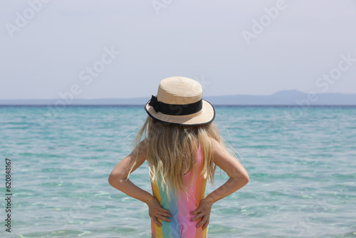 Outdoor portrait of little girl wearing straw hat on the beach and looking at beautiful blue sea horizon, summer vacation concept