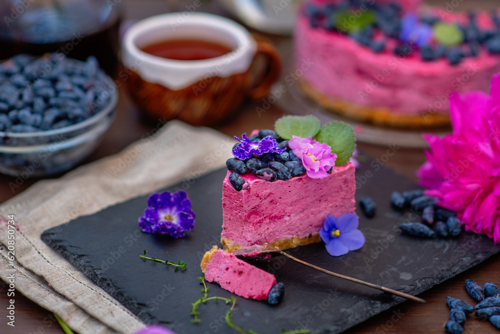 A piece of berry cheesecake lying next to a mug of tea, violet flowers and on a background of peonies and a whole cake A piece of berry cheesecake lying next to a mug of tea, violet flowers and on a background of peonies and a whole cake