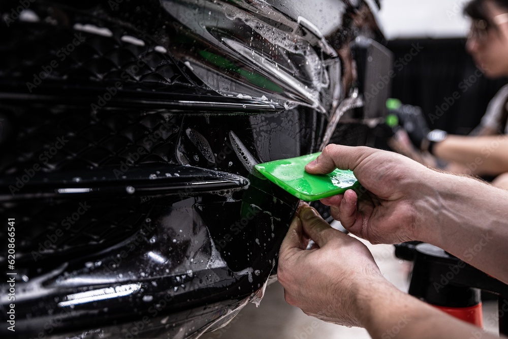 Car detailing studio employee, a wrapper, is protecting the bumper of a black car with a colorless protective PPF film. The PPF film protects the paint from scratches and damage.