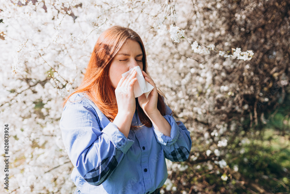 Sneezing young redhead woman with nose wiper among blooming trees in park. Portrait of sick women sneezes in white tissue, suffers from rhinitis and running nose. Symptoms of cold or allergy. - obrazy, fototapety, plakaty 