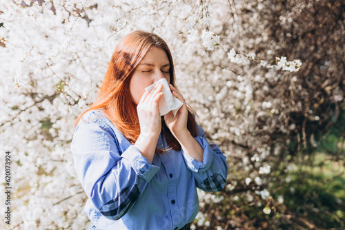 Sneezing young redhead woman with nose wiper among blooming trees in park. Portrait of sick women sneezes in white tissue, suffers from rhinitis and running nose. Symptoms of cold or allergy. photo
