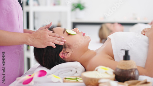 Beautiful woman lying on spa bed getting masking eyes with cucumber slices and relaxing with facial treatment by therapist. Skin care concept 
