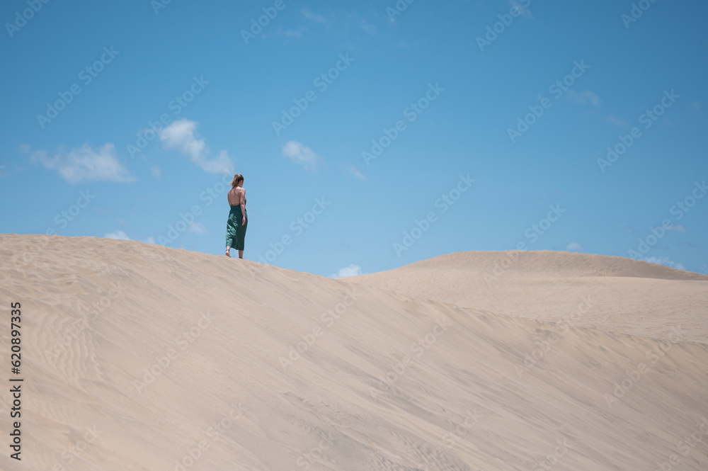 A blonde young girl in a green dress in the distance walking along the crest of the dune