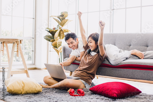 Happy Asian couple shopping online with laptop computer at home. Lovely couple making purchases online or booking in beautiful living room. Buying ordering products on the internet.