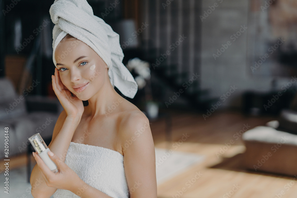 Skin care concept. Woman applies face lotion, wrapped in towel. Daily pampering, facial treatments. Bare shoulders, healthy complexion. Skin care concept. Woman applies face lotion, wrapped in towel. Daily pampering, facial treatments. Bare shoulders, healthy complexion.