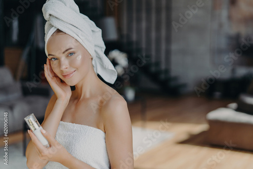 Skin care concept. Woman applies face lotion, wrapped in towel. Daily pampering, facial treatments. Bare shoulders, healthy complexion.