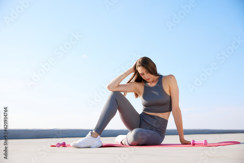 Young woman with slim, fit body sitting on fitness mantra in comfortable sportswear in sunny morning. Outdoor training