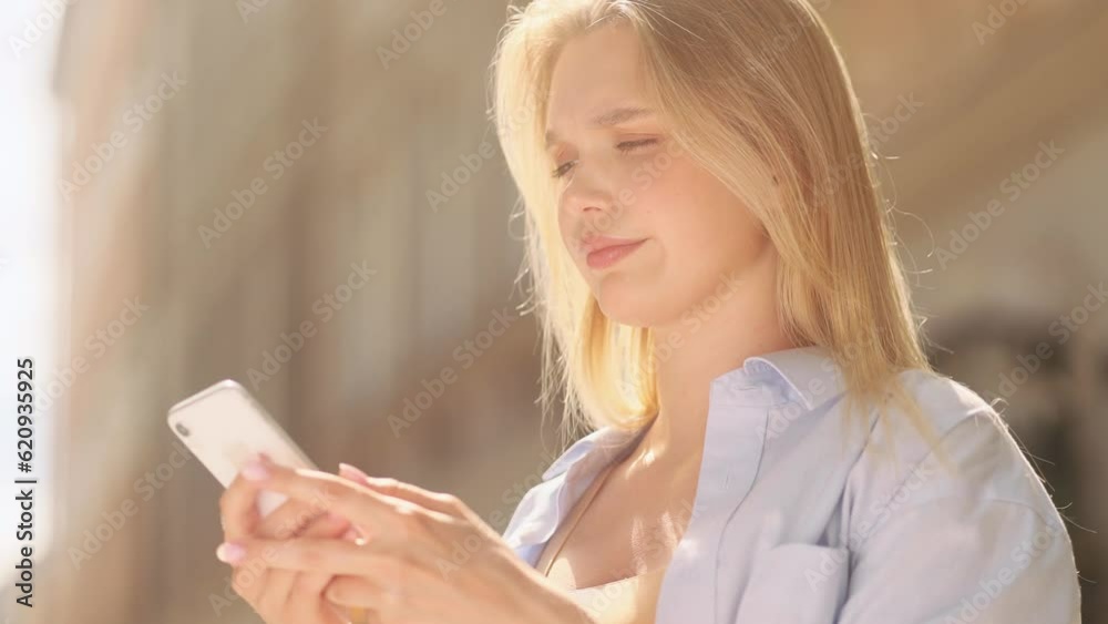 Irritated young blond woman texting at mobile phone at city centre Nervous female typing on smartphone expressing negative emotion outdoors Bad conflict working or personal conversation Fraud