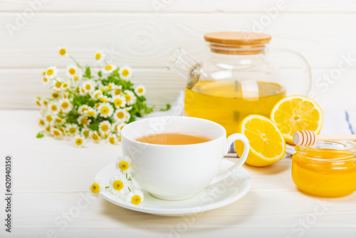 Fototapeta Naklejka Na Ścianę i Meble -  Chamomile herbal tea with flower buds, honey and lemon on a white wooden table and a bouquet of chamomile. Useful herbal, soothing drinks and natural healer concept. Immunity tea.Close up. Copy space.