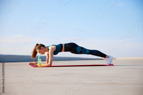 Sportive young woman in comfortable leggings and sport top standing in plank pose, training outdoors on warm summer day