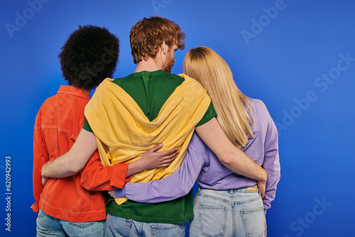 love triangle, polyamory lovers, back view of young redhead man hugging multiracial women on blue background, studio shot, vibrant colors, casual clothes, stylish attire, modern family photo