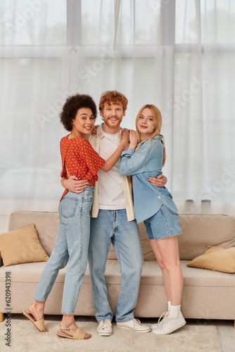 polygamy and love concept, three adults, happy redhead man hugging with multicultural women, threesome, cultural diversity, acceptance, bonding and love, multiracial lovers in living room © LIGHTFIELD STUDIOS