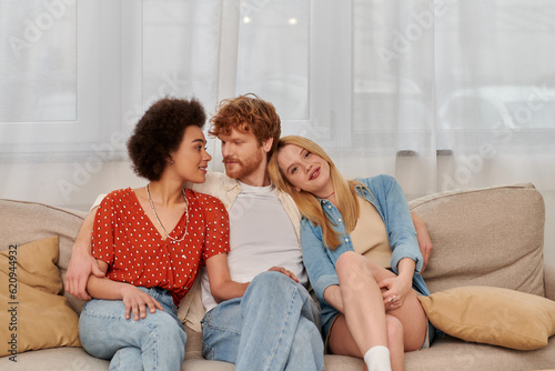 modern family, polygamy concept, freedom in relationship, cultural diversity, redhead man sitting with multicultural women on couch in living room, polyamorous lifestyle, non traditional photo