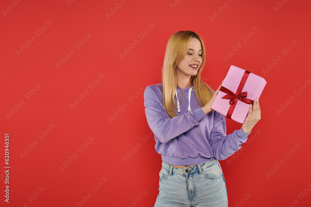 holiday, cheerfulness, blonde woman in casual attire holding present on coral background, smiling, vibrant colors, wrapped gift box, attractive and stylish, festive occasions holiday, cheerfulness, blonde woman in casual attire holding present on coral background, smiling, vibrant colors, wrapped gift box, attractive and stylish, festive occasions