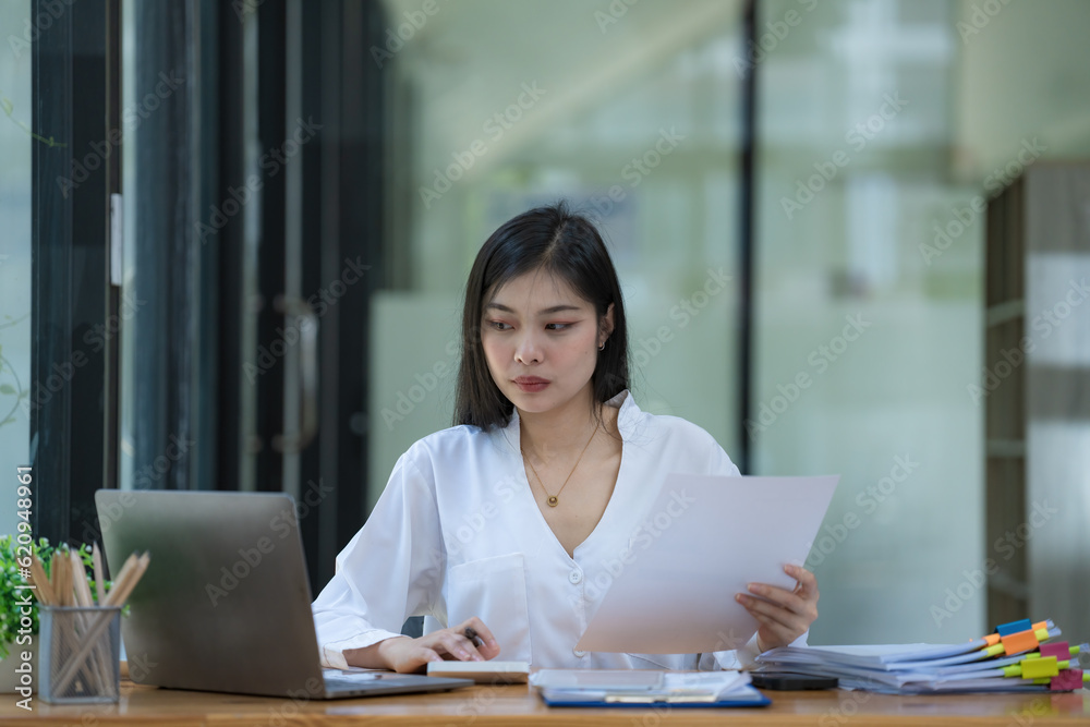 Thinking Asian businesswoman looking at financial statements and making marketing plans using a computer on her desk in the office holding report papers girl reading financial information. Thinking Asian businesswoman looking at financial statements and making marketing plans using a computer on her desk in the office holding report papers girl reading financial information.
