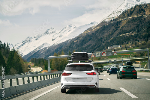 Rear view of a car with a roof box. Black luggage compartment box on a sport white car, back view. Alpine highway. Black Roof Box on a Sporty White Wagon Family Car © AlexGo