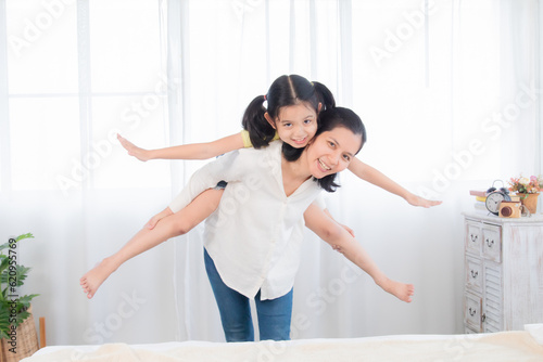 Cute little Asian girl piggybacks single mother having fun at home, Adorable daughter rides on mother with fun and gets ready to fly. Mom and child spend time together on holidays.