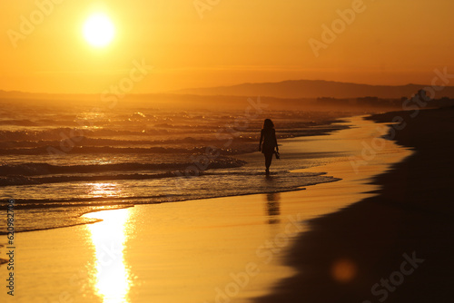A serenidade do entardecer na praia do Algarve, em Portugal. Uma mulher caminha descalça em direção ao horizonte, aproveitando o verão a beira mar. O cenário idílico proporciona um momento de paz.