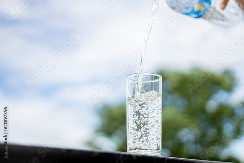 Fresh drinking water from bottle pouring into glass on table with blurred green nature background.