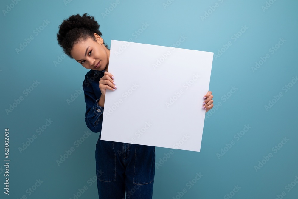 young brunette latin woman dressed in blue denim overalls holding a white board with a mockup for advertising