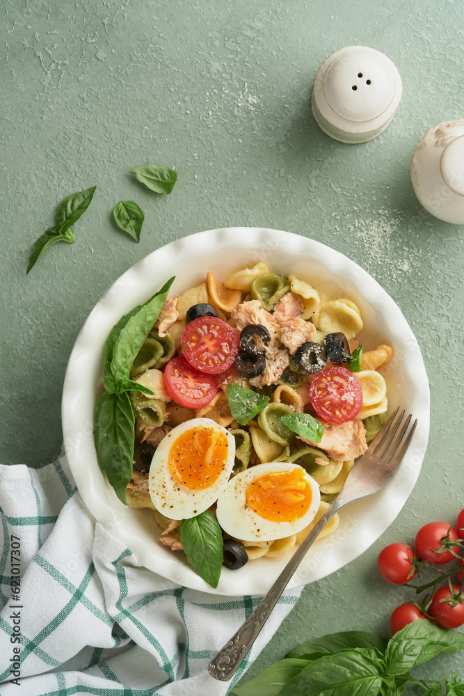 Italian pasta salad. Orecchiette pasta with tuna, tomato cherry, olive, basil and parmesan cheese in plate on green stone or concrete background. Traditional Italian cuisine. Flat lay. Copy space.