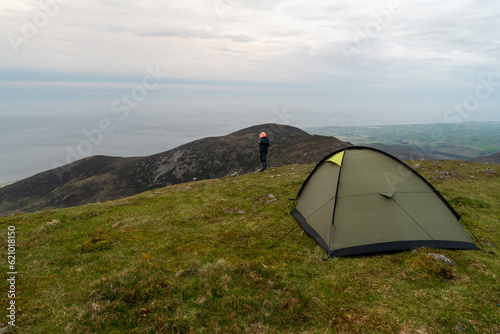 Green tent in the mountains under evening sky. Woman hiker enjoying the view.  Typical Irish mountains  landscape. photo