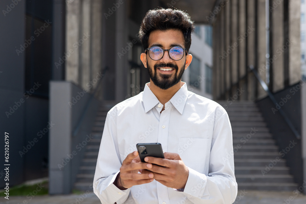 Portrait of young indian programmer businessman, man in glasses holding phone, looking at camera and smiling, happy with achievement results freelancer. Portrait of young indian programmer businessman, man in glasses holding phone, looking at camera and smiling, happy with achievement results freelancer.