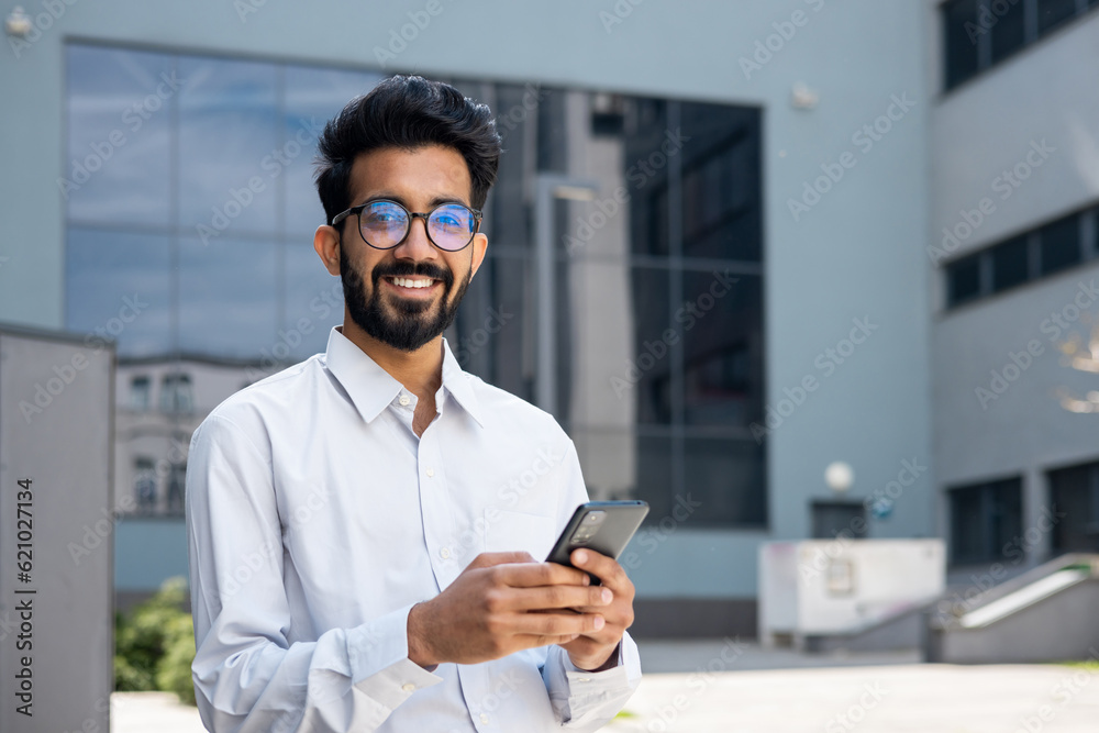 Portrait of young indian programmer businessman, man in glasses holding phone, looking at camera and smiling, happy with achievement results freelancer. Portrait of young indian programmer businessman, man in glasses holding phone, looking at camera and smiling, happy with achievement results freelancer.