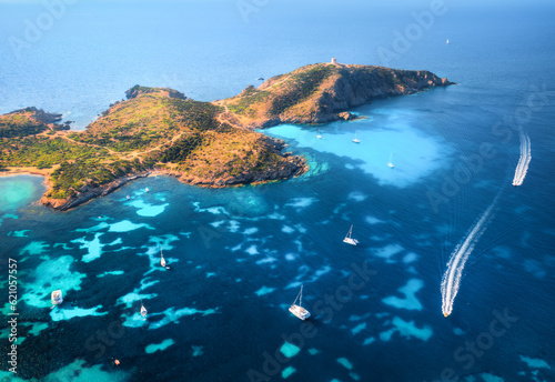 Aerial view of island, sea bay with clear azure water, yachts and boats on blue sea at sunset in summer. Sardinia, Italy. Tropical seascape with speed boats, mountain, ocean. Top drone view