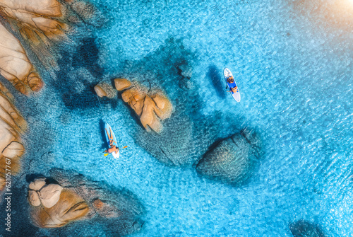 Aerial view of sup boards, blue sea, rocks at sunrise in summer. People on floating canoe in transparent azure water. Kayak. Sardinia island, Italy. Tropical seascape. Active travel. Top drone view
