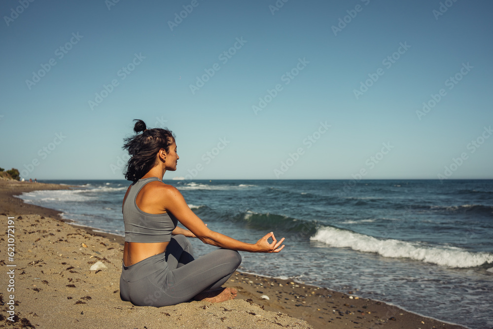 a beautiful athletic brunette girl in a gray tracksuit is engaged in meditation sitting on the sand against the backdrop of the sea