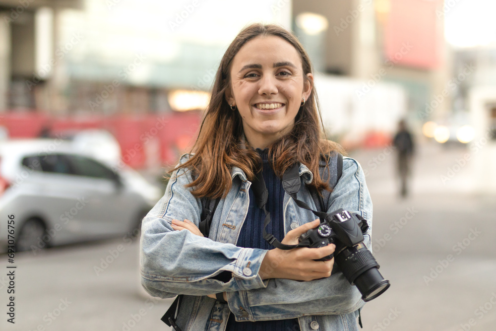 Young woman in casual attire with professional camera in hand smiling. Blurred city background.