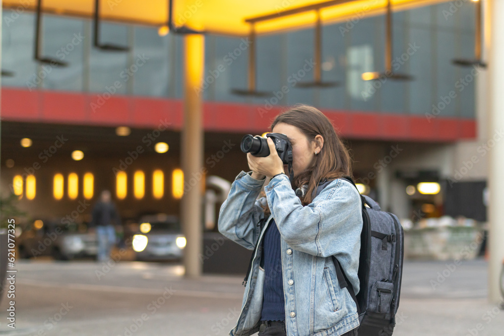 Photographer with professional camera covering her face taking photo in the city. Colorful blurred background. Contrasts.