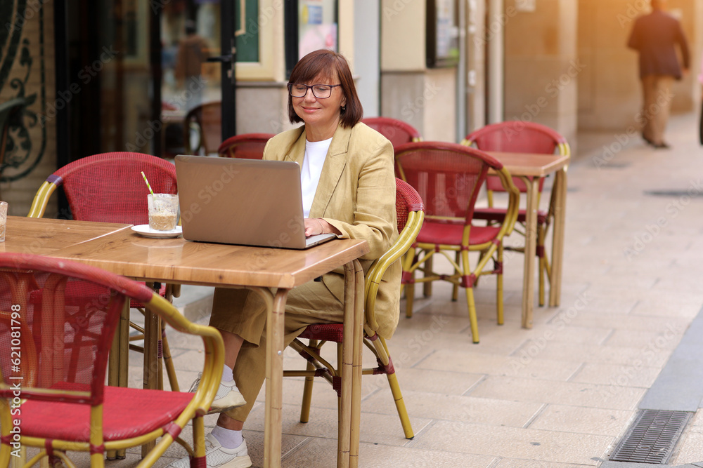 Fototapeta premium Mature stylish businesswoman in eyeglasses works on laptop while sitting with a coffee drink at European cafe outdoors. Concept of remote work from public place, digital freelance and modern lifestyle