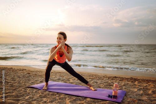 Beautiful young woman in sportswear practicing stretching on yoga mat alone on wild beach. Healthy lifestyle concept